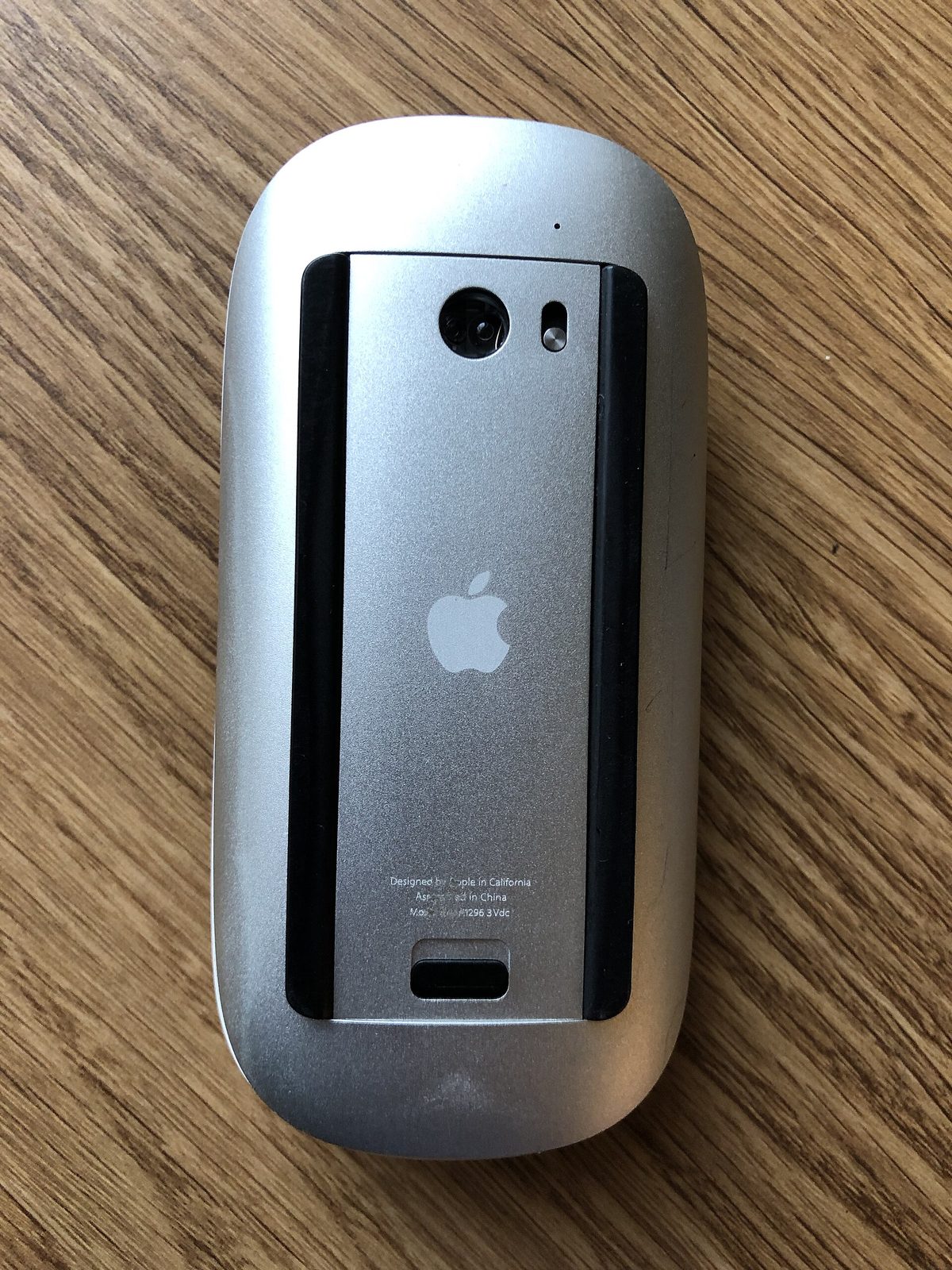 Underside of a Magic Mouse v1. A wide silver battery door with a release latch covers the whole middle of the bottom. There is no charging port.